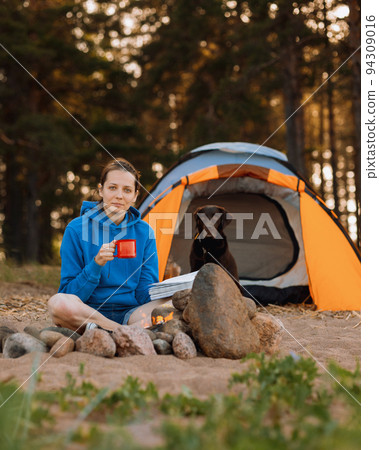 woman and a Labrador retriever dog on a camping trip with a tent in nature. a woman is reading a book or drinking tea from a red cup next to a campfire 94309016