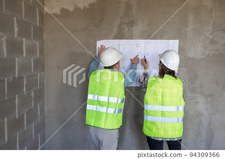 two engineers, a man and a woman in white helmets and protective vests, are standing in the room and holding a plan of the construction two engineers, a man and a woman in white helmets and protective vests, are standing in the room and holding a plan of the construction 94309366