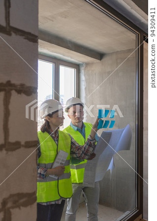 two engineers in protective vests and white helmets glue stickers on the window and discuss the construction plan of the facility or the scheme 94309411