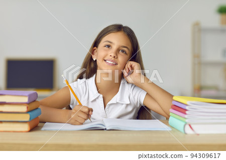 Portrait of funny and cute schoolgirl with dreamy expression sitting at desk between books. 94309617