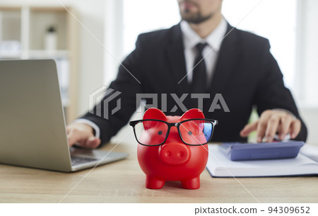 Close up of red piggy bank in form of pig stands on table against male financier. Close up of red piggy bank in form of pig stands on table against male financier. 94309652