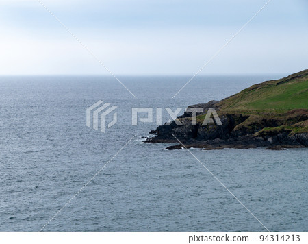 View of the coastal cliffs from the Warren beach. The rocky coast of the south of Ireland. Hills under the sky on a spring day, mountain beside sea under white clouds and sky. View of the coastal cliffs from the Warren beach. The rocky coast of the south of Ireland. Hills under the sky on a spring day, mountain beside sea under white clouds and sky. 94314213