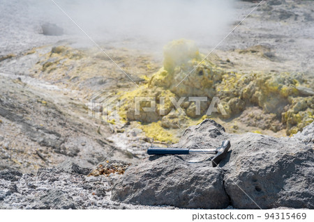 geological hammer on the rock against the backdrop of an steaming fumarole on the slope of a volcano geological hammer on the rock against the backdrop of an steaming fumarole on the slope of a volcano 94315469