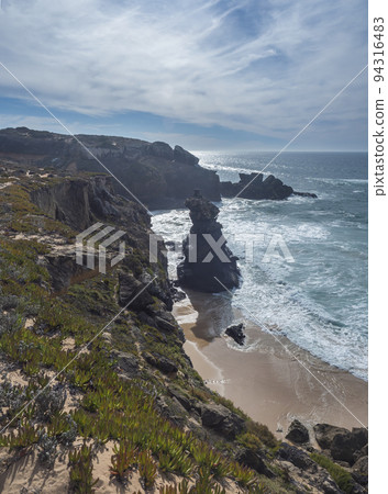 View of sharp cliffs and small sand beach with ocean waves, rocks, stones and green vegetation at wild Rota Vicentina coast near Vila Nova de Milfontes, Portugal. 94316483