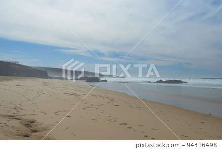 View of Praia dos Aivados sand beach with ocean waves and sharp rock and distant figures at wild Rota Vicentina coast near Porto Covo, Portugal. View of Praia dos Aivados sand beach with ocean waves and sharp rock and distant figures at wild Rota Vicentina coast near Porto Covo, Portugal. 94316498