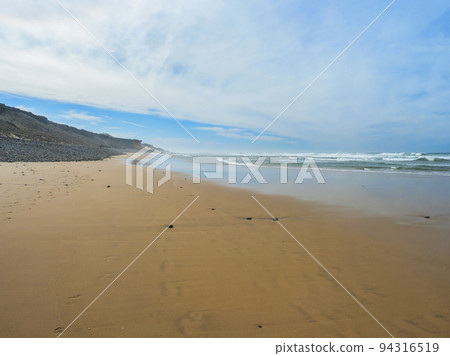 View of empty Praia do Queimado beach with ocean waves and sharp rocks and wet golden sand at wild Rota Vicentina coast near Porto Covo, Portugal. 94316519