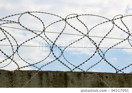 Barbed wire on the fence. Fencing stainless steel Barb Wire with sharp edges on a massive concrete wall for security on the background a blue sky. Barbwire on the prison fence. Military barbwire fence Barbed wire on the fence. Fencing stainless steel Barb Wire with sharp edges on a massive concrete wall for security on the background a blue sky. Barbwire on the prison fence. Military barbwire fence 94317091