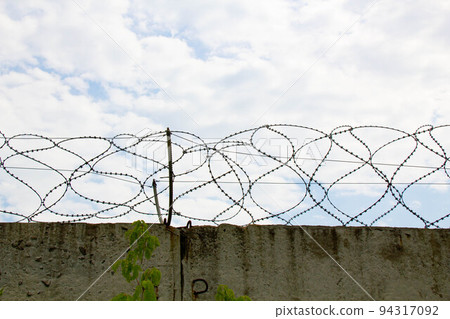 Barbed wire on the fence. Fencing stainless steel Barb Wire with sharp edges on a massive concrete wall for security on the background a blue sky. Barbwire on the prison fence. Military barbwire fence Barbed wire on the fence. Fencing stainless steel Barb Wire with sharp edges on a massive concrete wall for security on the background a blue sky. Barbwire on the prison fence. Military barbwire fence 94317092