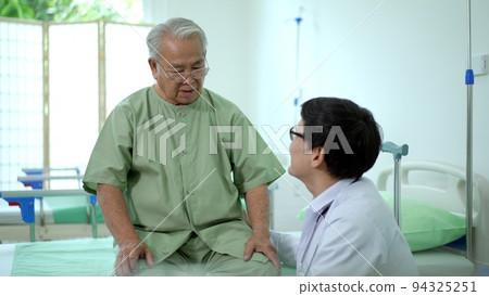 Elder Asian man sitting on the bed and talking to doctor about his physical injuries at his knees, elderly Asian man having a consulting with professional physical therapist or orthopedic doctor. 94325251