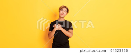 Portrait of touched handsome asian guy holding hands on heart and smiling flattered, yellow background 94325536