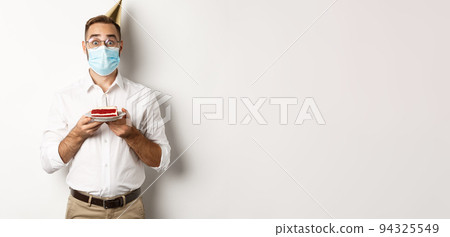 Covid-19, social distancing and celebration. Surprised birthday guy holding bday cake, wearing face mask from coronavirus, white background 94325549