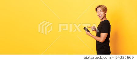 Portrait of pleased smiling handsome asian man, standing in profile and pointing finger at smartphone, recommend app, standing yellow background 94325669