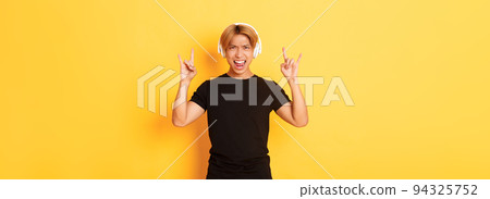 Joyful handsome asian guy enjoying listening music in headphones, showing rock-n-roll gesture, standing over yellow background Joyful handsome asian guy enjoying listening music in headphones, showing rock-n-roll gesture, standing over yellow background 94325752
