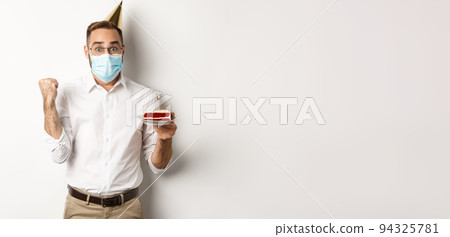 Covid-19, social distancing and celebration. Hopeful happy birthday man in face mask, holding bday cake and rejoicing, standing against white background 94325781