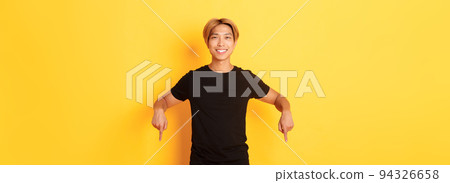 Portrait of smiling attractive korean guy with blond hair, wearing black outfit, pointing fingers down, showing way, standing yellow background Portrait of smiling attractive korean guy with blond hair, wearing black outfit, pointing fingers down, showing way, standing yellow background 94326658