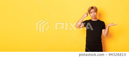 Disappointed asian guy with blond hair, raising hand up puzzled and scolding someone acting stupid, standing yellow background 94326813