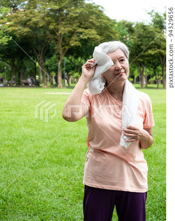 快樂的樂齡亞洲婦女微笑著站在草地上,脖子披著運動毛巾,享受公園裡的大自然新鮮空氣. 快樂的樂齡亞洲婦女微笑著站在草地上,脖子披著運動毛巾,享受公園裡的大自然新鮮空氣. 94329656