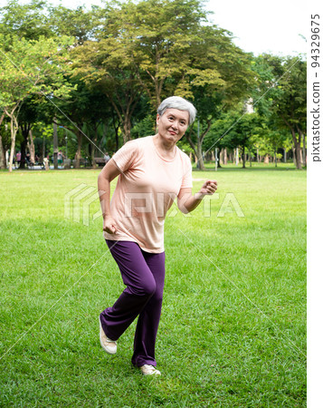 Happy senior Asian woman smiling and jogging on grass and enjoying nature fresh air in park. 94329675