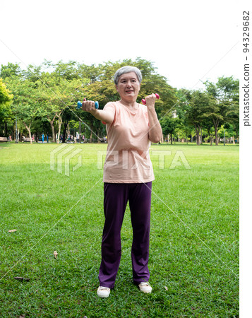 快樂的樂齡亞洲婦女微笑著站在草地上使用啞鈴運動,並且享受公園裡的大自然新鮮空氣. 快樂的樂齡亞洲婦女微笑著站在草地上使用啞鈴運動,並且享受公園裡的大自然新鮮空氣. 94329682