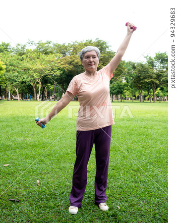 快樂的樂齡亞洲婦女微笑著站在草地上使用啞鈴運動,並且享受公園裡的大自然新鮮空氣. 快樂的樂齡亞洲婦女微笑著站在草地上使用啞鈴運動,並且享受公園裡的大自然新鮮空氣. 94329683