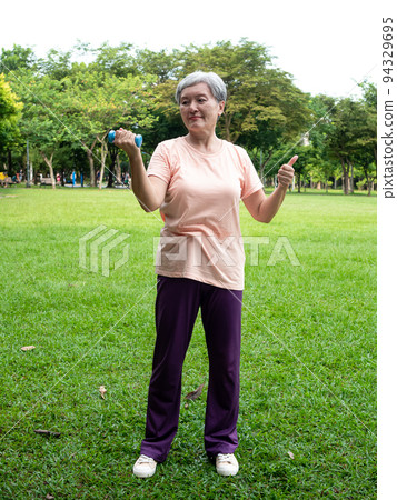 快樂的樂齡亞洲婦女微笑著站在草地上使用啞鈴運動,並且享受公園裡的大自然新鮮空氣. 快樂的樂齡亞洲婦女微笑著站在草地上使用啞鈴運動,並且享受公園裡的大自然新鮮空氣. 94329695