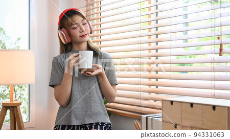 Calm young woman holding cup of coffee and listening yo music on wireless headphone, enjoy stress free peaceful mood Calm young woman holding cup of coffee and listening yo music on wireless headphone, enjoy stress free peaceful mood 94331063