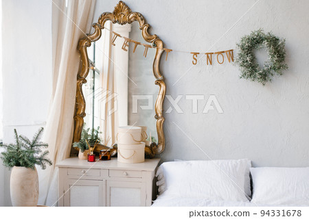 The interior of a bedroom decorated for Christmas and New Year in a Scandinavian or vintage style. A mirror over the dresser, spruce branches in a vase, a Christmas wreath over the bed 94331678