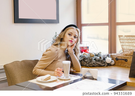 An attractive happy girl is sitting in a cafe decorated for Christmas, drinking coffee and eating dessert 94331689