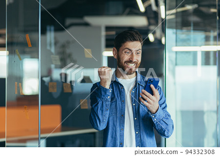 Excited happy business man with a beard, a professional winner, celebrating success by reading good news on a smartphone, feeling excited about mobile messaging Excited happy business man with a beard, a professional winner, celebrating success by reading good news on a smartphone, feeling excited about mobile messaging 94332308