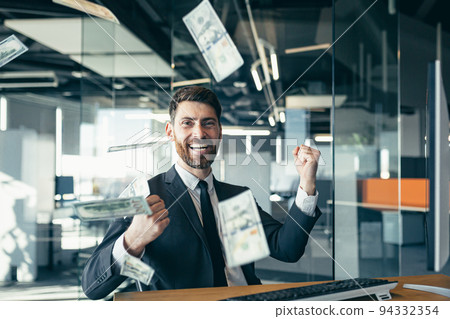 Handsome joyful young businessman sitting at a desk, working in a modern office, throwing money over his head. Happy smiling man throws money in the air Handsome joyful young businessman sitting at a desk, working in a modern office, throwing money over his head. Happy smiling man throws money in the air 94332354