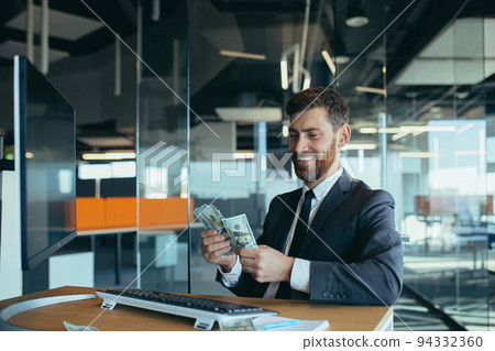 Joyful business man in sitting at desk working on laptop computer in bright office Achieving business career concept. Keep cash by making a gesture to the winner Joyful business man in sitting at desk working on laptop computer in bright office Achieving business career concept. Keep cash by making a gesture to the winner 94332360