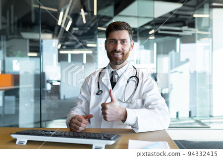 Successful doctor works sitting at the table looks at the camera and smiles makes a positive gesture with his hands happy, works in a modern clinic 94332415