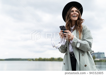 portrait of a smiling young woman in a black hat and autumn gray coat with a phone on the background portrait of a smiling young woman in a black hat and autumn gray coat with a phone on the background 94332805