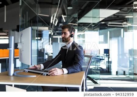 Business man in a modern office works at a computer, uses a headset for video calling, technical support Business man in a modern office works at a computer, uses a headset for video calling, technical support 94332959