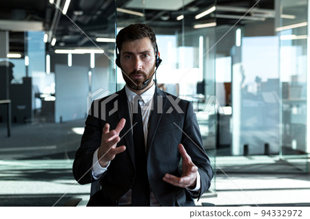 The employee makes a video call, looks at the camera and explains the task, serious gestures with his hands works in a modern office, uses a headset The employee makes a video call, looks at the camera and explains the task, serious gestures with his hands works in a modern office, uses a headset 94332972