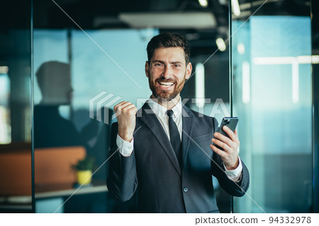 Happy businessman boss looks at camera and smiles, holds phone in hands, man wins online and celebrates victory Happy businessman boss looks at camera and smiles, holds phone in hands, man wins online and celebrates victory 94332978