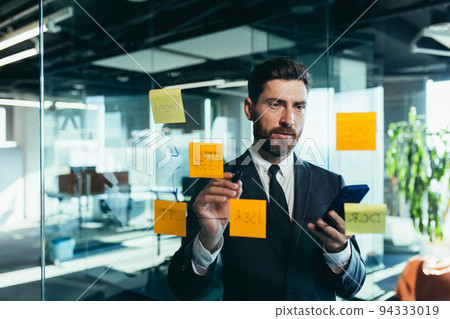 Successful businessman rewrites a task on a glass board with colored stickers, from phone and tablet Successful businessman rewrites a task on a glass board with colored stickers, from phone and tablet 94333019
