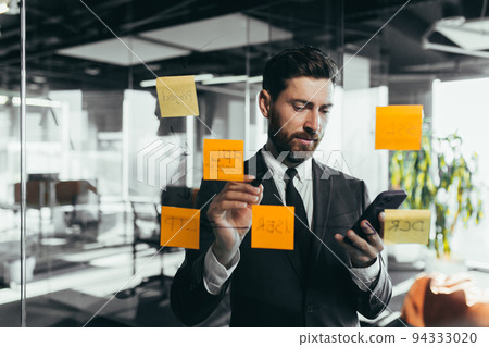 Successful businessman rewrites a task on a glass board with colored stickers, from phone and tablet Successful businessman rewrites a task on a glass board with colored stickers, from phone and tablet 94333020