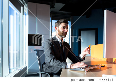 cheerful bearded businessman shopping online working in a modern office computer, man holding a bank credit card cheerful bearded businessman shopping online working in a modern office computer, man holding a bank credit card 94333141