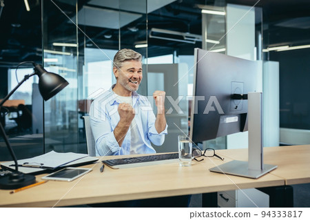 Experienced gray-haired businessman celebrating victory working at computer in modern office at computer, man looking at camera and smiling hand gesture up Experienced gray-haired businessman celebrating victory working at computer in modern office at computer, man looking at camera and smiling hand gesture up 94333817