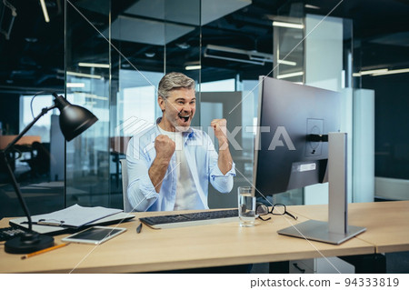Experienced gray-haired businessman celebrating victory working at computer in modern office at computer, man looking at camera and smiling hand gesture up 94333819