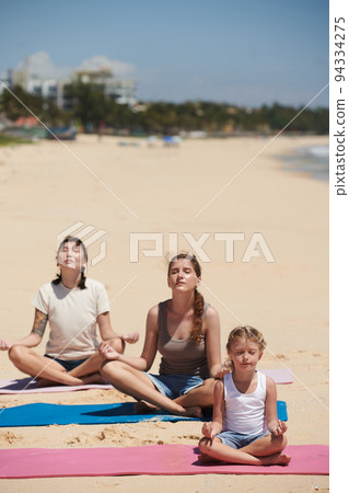 Little girl and two women meditating on beach on sunny morning 94334275