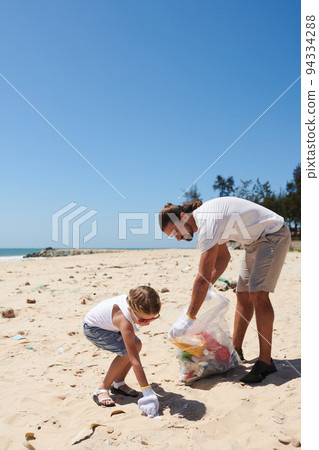 Little girl helping father to collect garbage on sea beach in big plastic bag 94334288