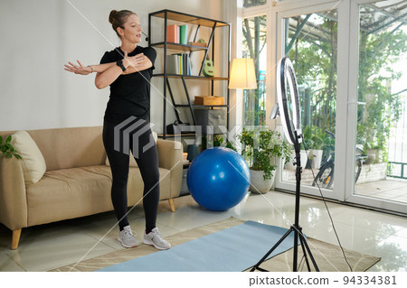 Fitness instructor recording video for her social media, explaining how to warm up before training Fitness instructor recording video for her social media, explaining how to warm up before training 94334381