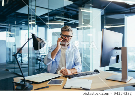 Gray-haired businessman with a runny nose working in the office at the computer, cold and flu Gray-haired businessman with a runny nose working in the office at the computer, cold and flu 94334382