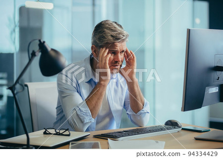 An experienced gray-haired businessman works at the computer until late, tired has a severe headache, stress and depression 94334429