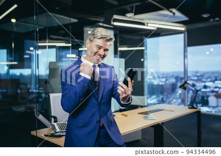 Satisfied and happy leader, senior and experienced businessman looks at the phone smiles and rejoices in victory, received good news 94334496