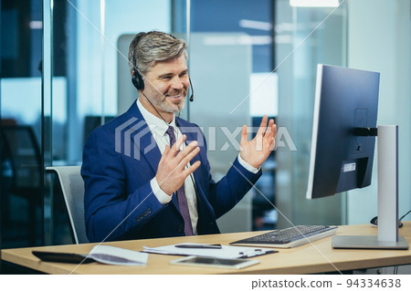 video communication man in a business suit looks at the webcam on a computer monitor, with a headset talks to colleagues online conference video communication man in a business suit looks at the webcam on a computer monitor, with a headset talks to colleagues online conference 94334638