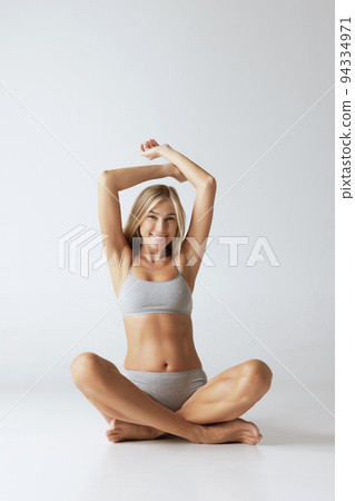 Young happy woman in grey cotton underwear sitting on floor and looking at camera witj smile isolated over gray background. Wellness, fitness, diet, relax 94334971