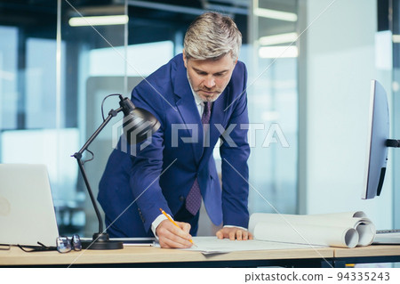Experienced architect, working at the office desk on the drawing, makes final edits before handing over the object, a man in a modern office Experienced architect, working at the office desk on the drawing, makes final edits before handing over the object, a man in a modern office 94335243
